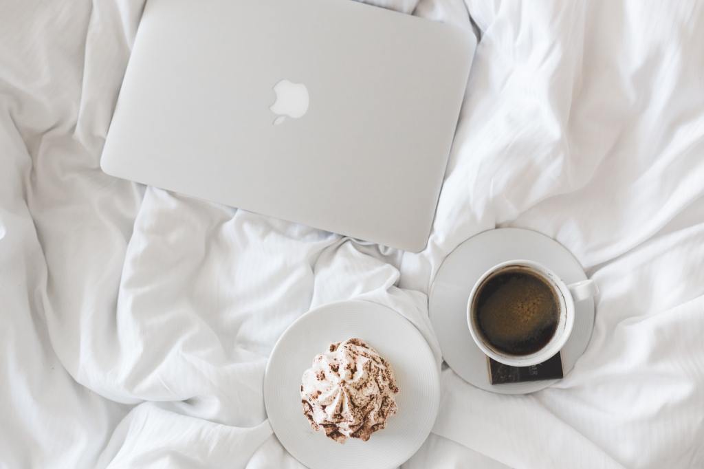 Cozy morning setup with white bed linens, closed silver MacBook, and breakfast tray with black coffee and a chocolate pastry. Inspires Christian self-care and mental health routines, including devotionals, Bible journaling, gratitude practices, and gentle faith-filled morning rituals.