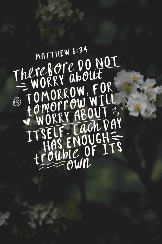 Hand-lettered Christian digital design featuring Matthew 6:34 Bible verse in elegant white calligraphy on a dark blurred floral background with soft white blossoms: “Therefore do not worry about tomorrow, for tomorrow will worry about itself. Each day has enough trouble of its own.” Inspirational Scripture artwork for overcoming anxiety with God through faith and trust in His daily provision.